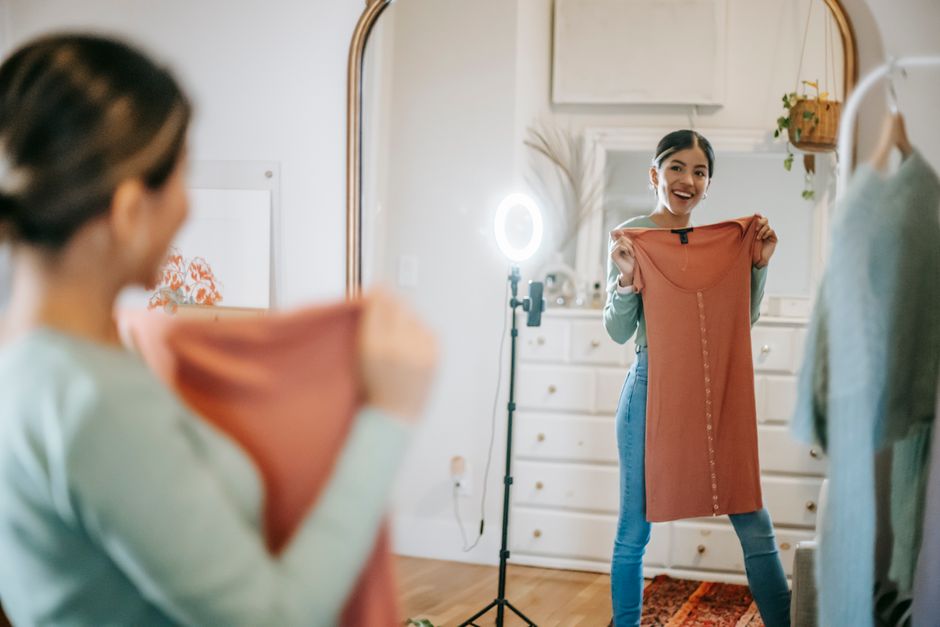 Smiling young ethnic female blogger in trendy outfit standing in room and demonstrating dress in hands while looking at mirror and shooting video on phone with ring lamp near cabinet