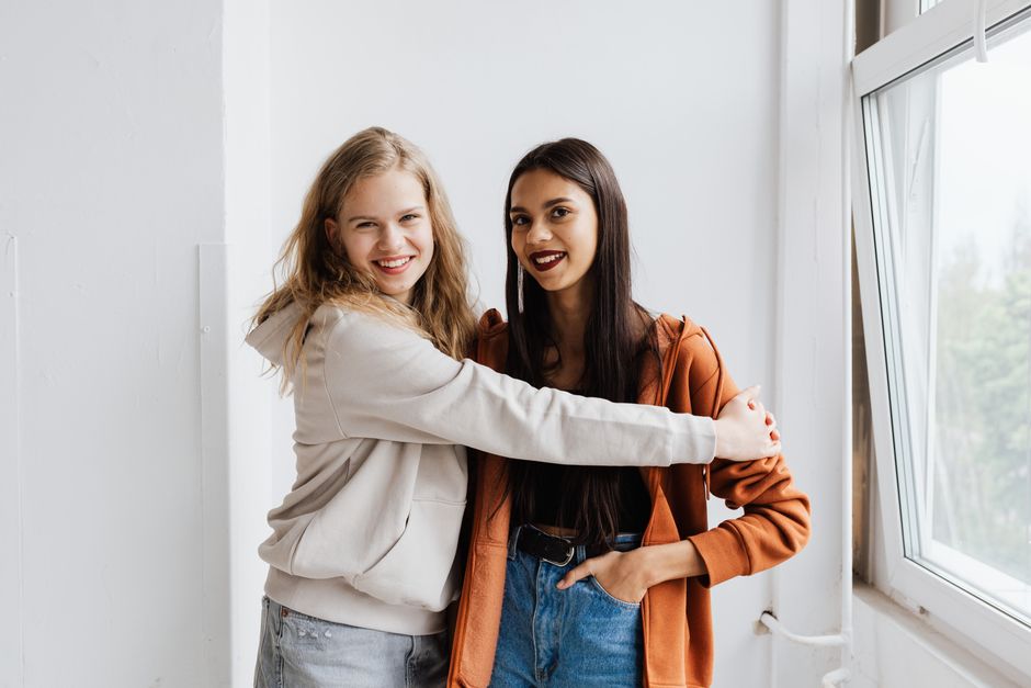Two women standing by a window, smiling and hugging, wearing casual hoodies.