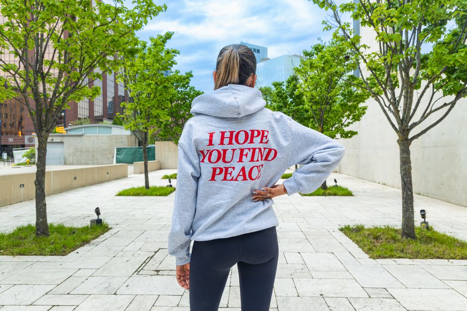 Back view of a woman in a gray hoodie with 'I Hope You Find Peace' text in a park setting.