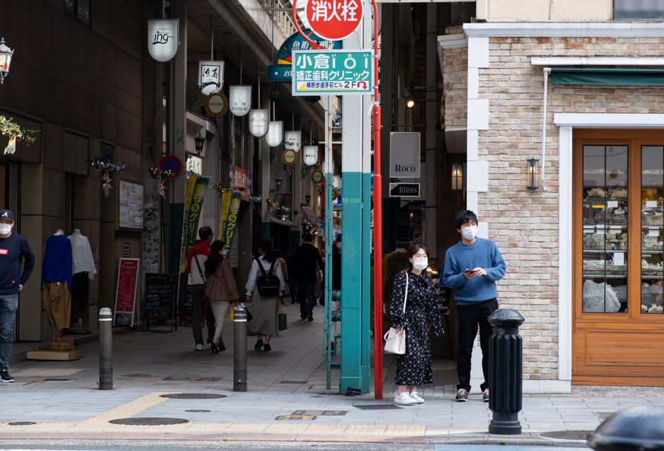 Urban street scene with people wearing masks, featuring shops and signage.