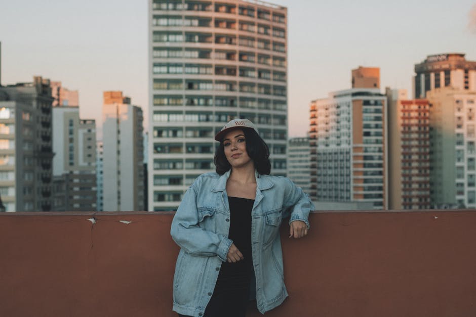 Woman posing on a rooftop with city skyline backdrop during twilight, wearing casual denim and cap.