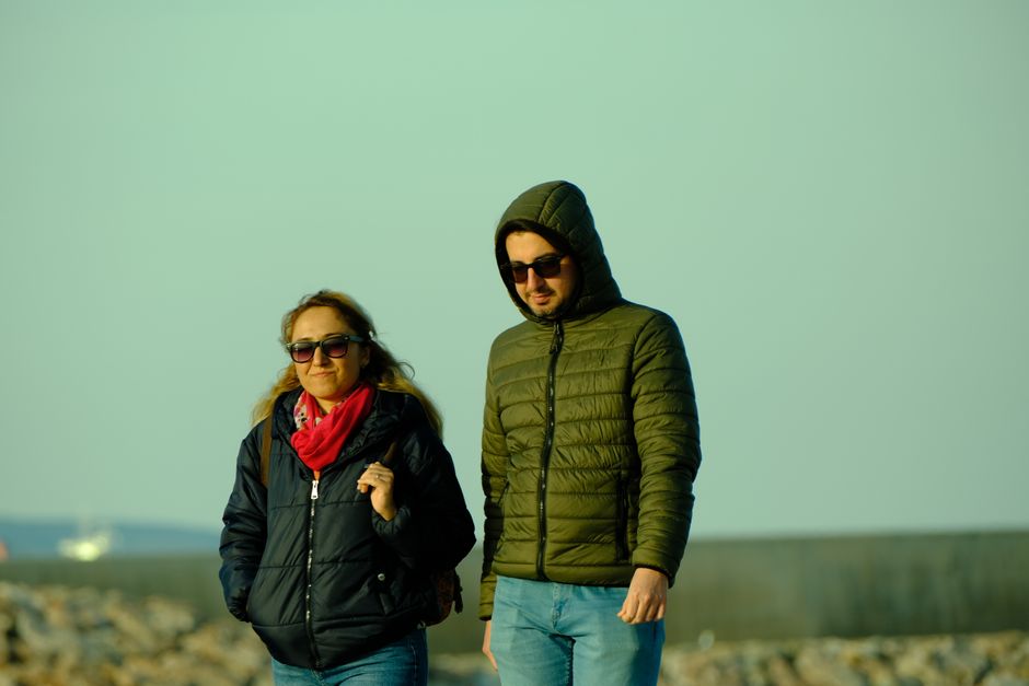 A couple wearing winter jackets and sunglasses walking on a rocky beach.