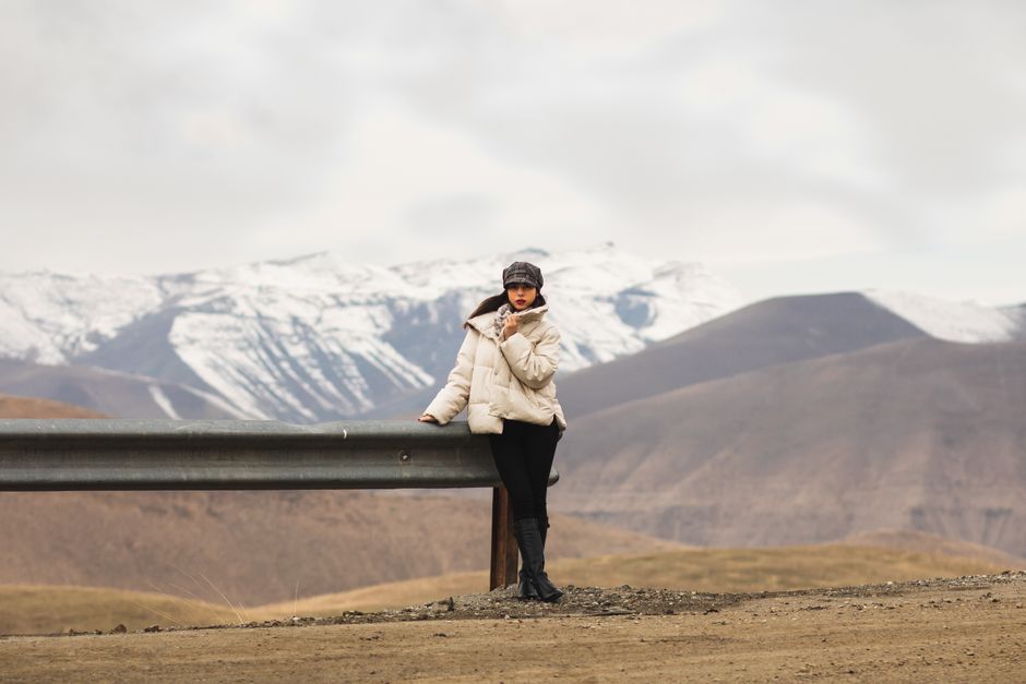 A woman in a puffer jacket poses by a mountain with snow-capped peaks.