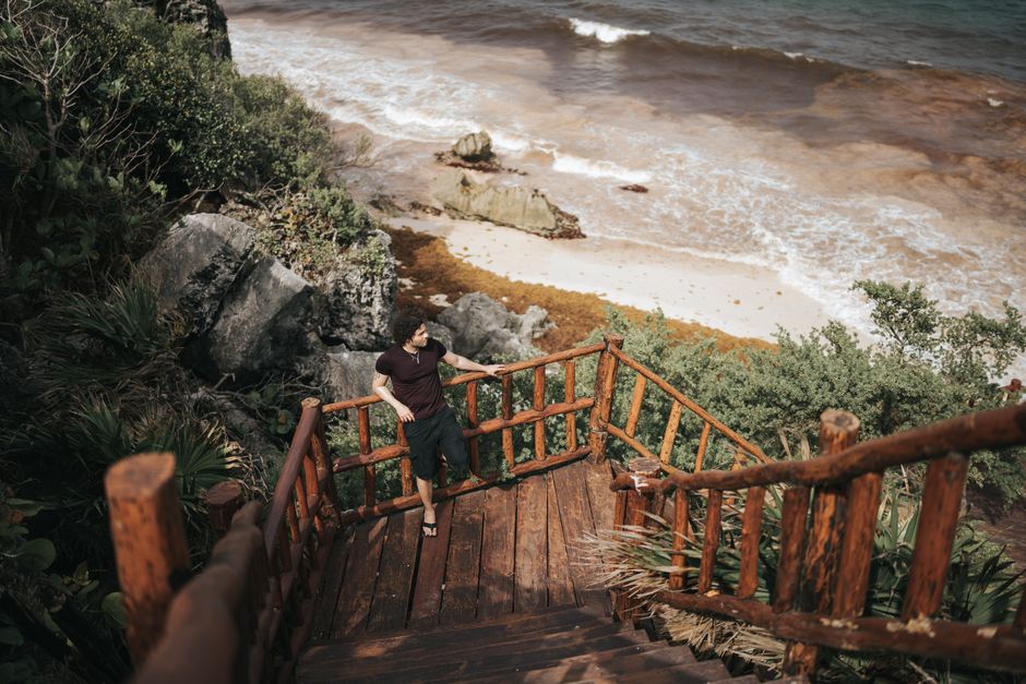 A man standing on wooden stairs overlooking a serene beach with lush greenery and rocky shore.