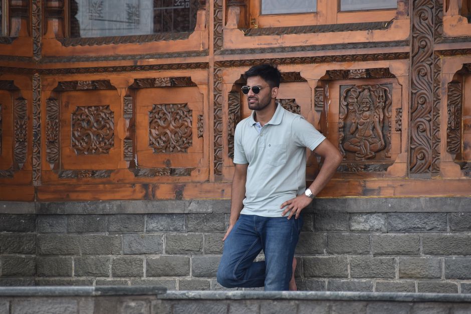 Man in sunglasses and casual outfit leaning against an ornate wooden wall outdoors.