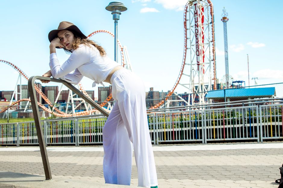 Fashionable woman poses at an amusement park with roller coaster backdrop, wearing casual attire and a hat.