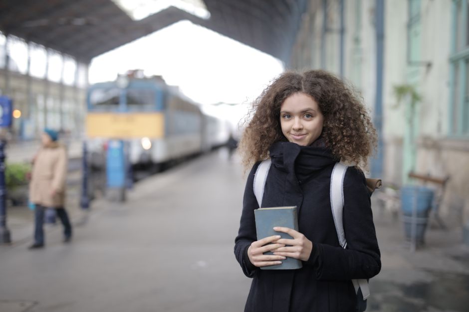 Positive young lady with curly hair wearing casual warm coat standing on platform in railway station with book and backpack and looking at camera