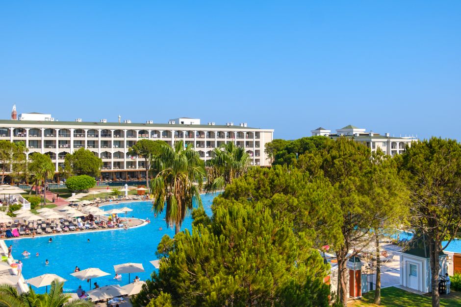 Aerial view of a resort with a large swimming pool surrounded by palm trees on a bright summer day.