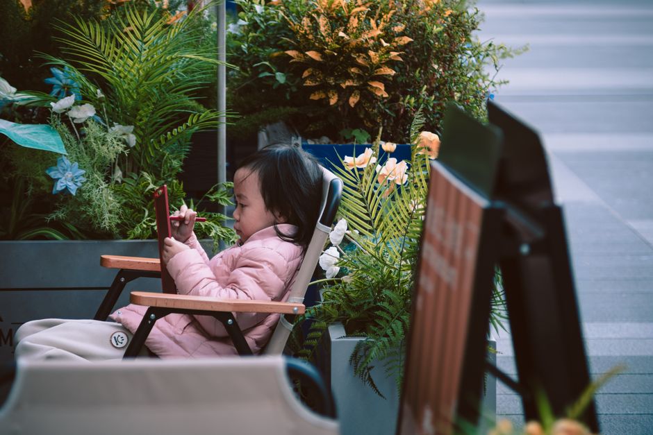 Young child sitting in a chair using a tablet, surrounded by vibrant plants and flowers outdoors.