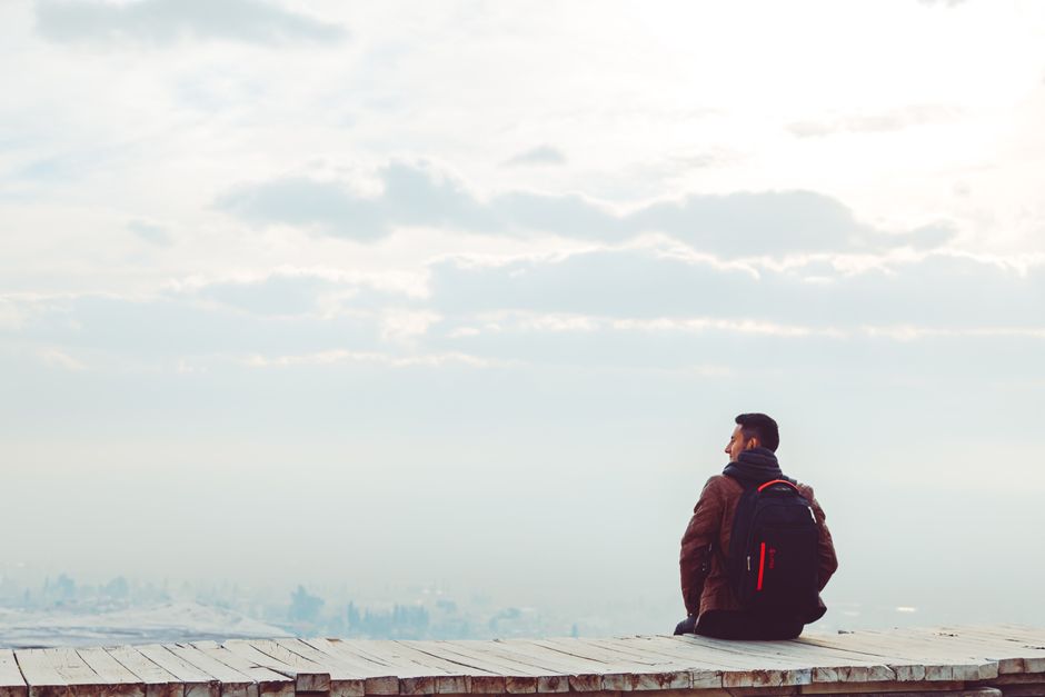A lone man with a backpack sits on a wooden platform under cloudy skies, embracing solitude.