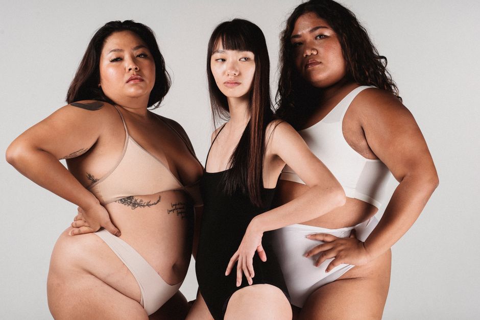 Unemotional Asian female in black bodysuit standing with hands on waist in light studio between plus size female models in white underwear