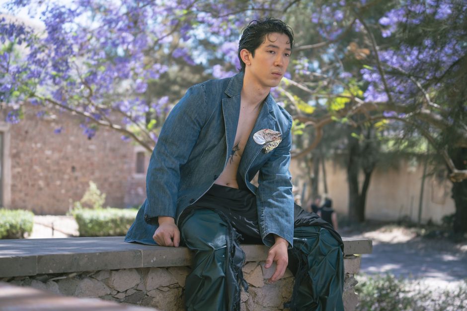 Young man in a denim jacket sits outdoors under blooming jacaranda trees, exuding a modern stylish vibe.