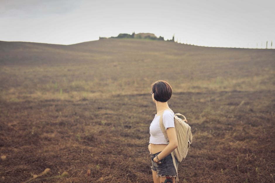 Woman with backpack stands in vast countryside field, evoking solitude.