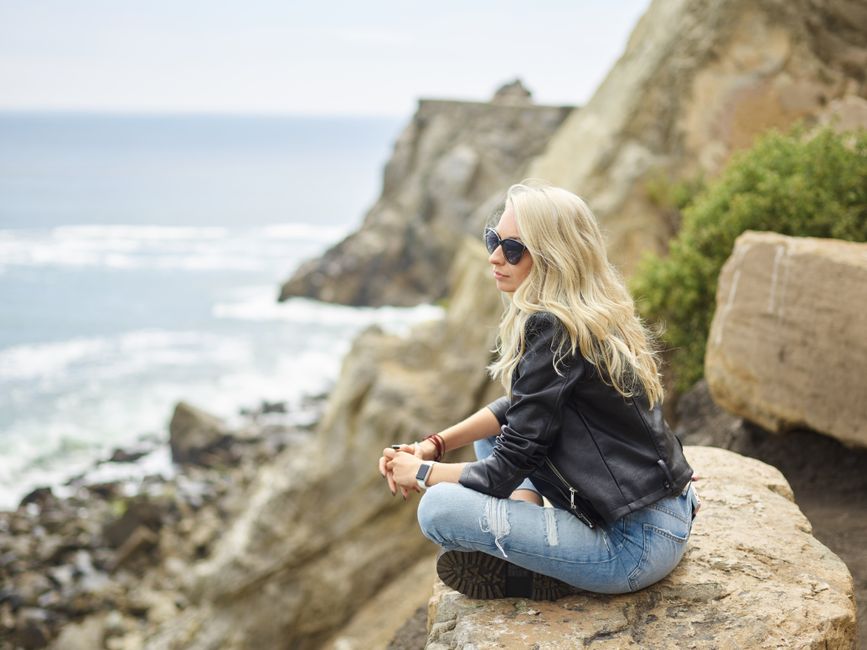 Side view of pensive young lady in leather jacket and sunglasses sitting on rocky cliff with crossed legs and admiring waving ocean