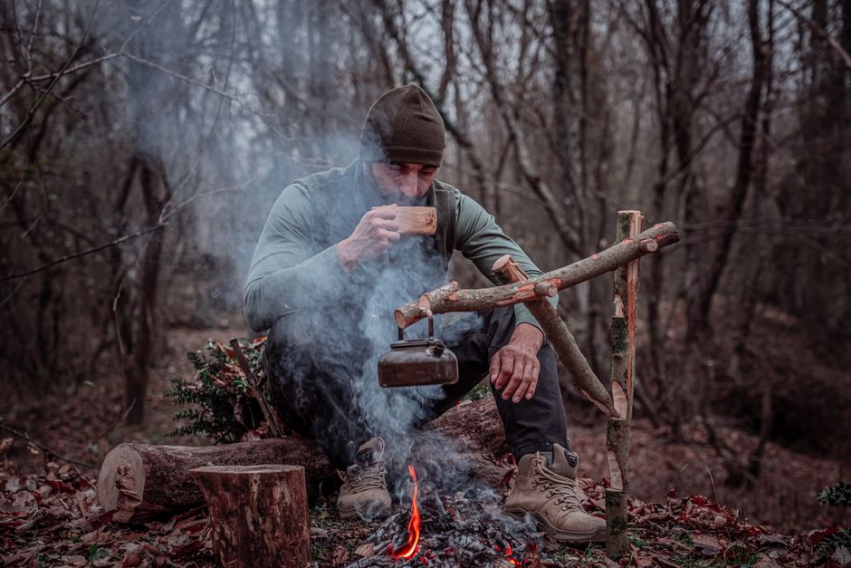 An adult man enjoys coffee by a campfire in a serene forest setting.