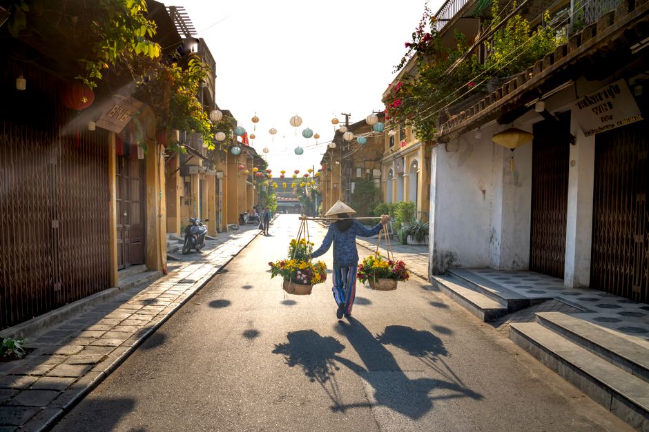 Back view of anonymous local woman in casual clothes and conical hat carrying heavy baskets of fresh flowers hanging on stick on neck while walking between residential houses in Vietnam