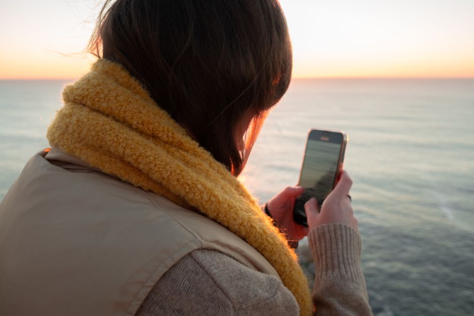 A woman taking a photo of the ocean sunset with her smartphone, wrapped in a warm scarf.