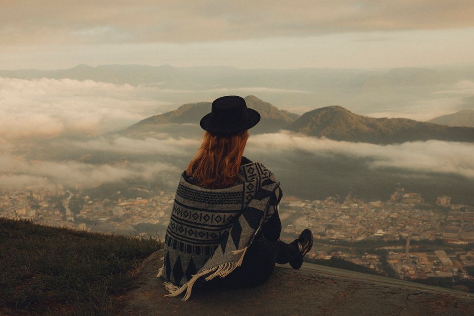 Young woman sitting on a mountaintop overlooking a foggy cityscape at dawn.