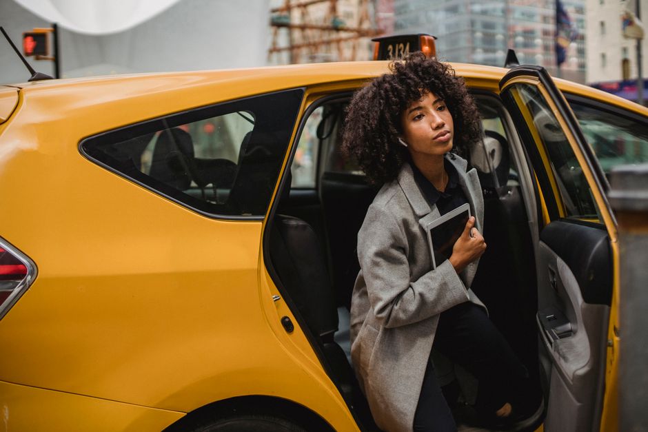 African American woman stepping out of a yellow taxi in a city setting, holding a tablet.