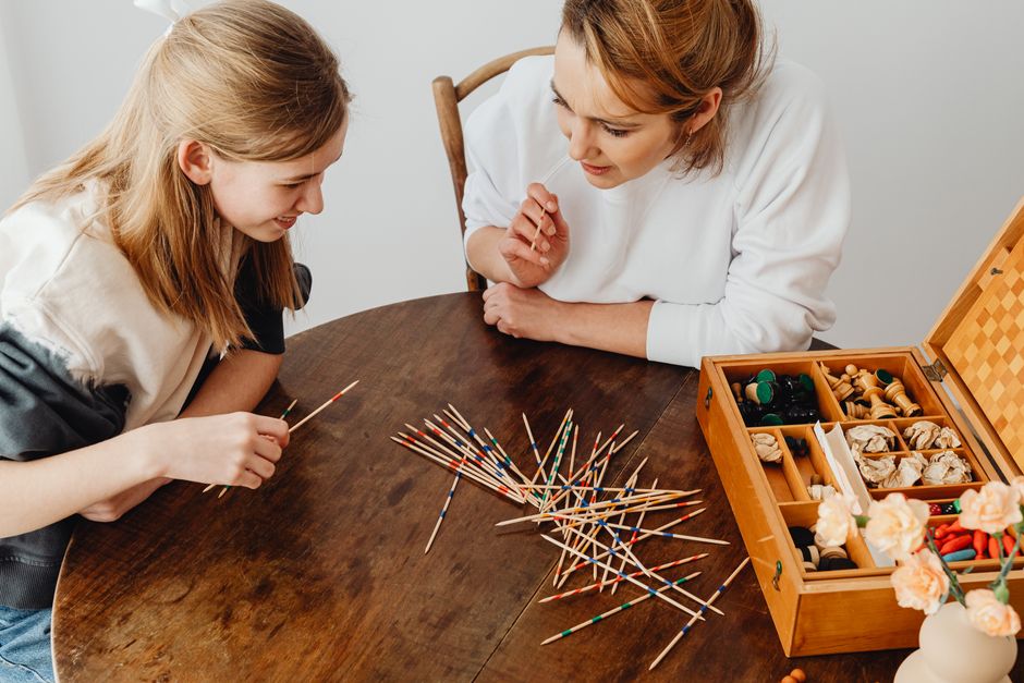 Two women enjoying a game of Mikado at a wooden table, engaging in conversation and play.