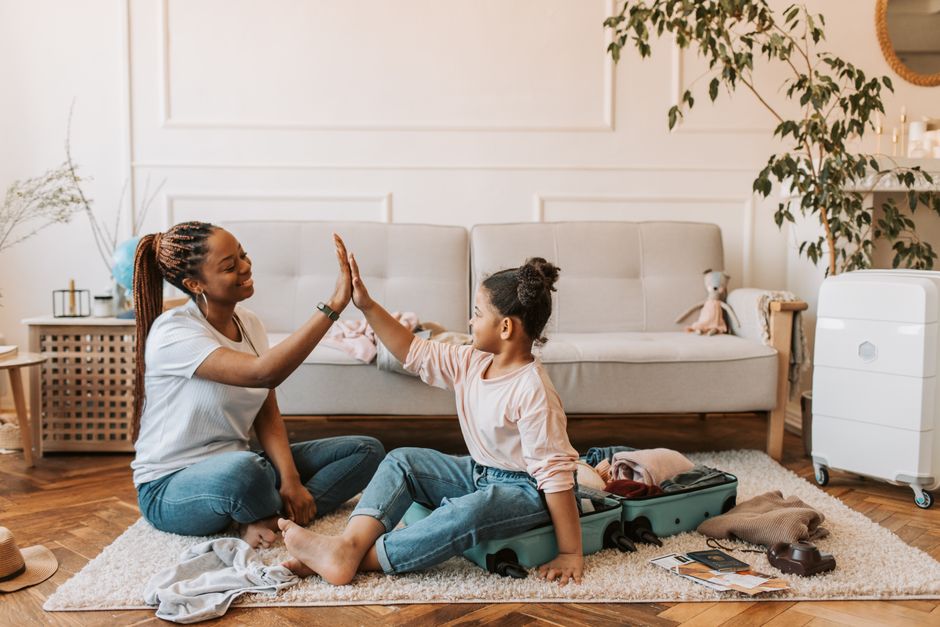 A mother and daughter high-five while packing a suitcase in their living room.